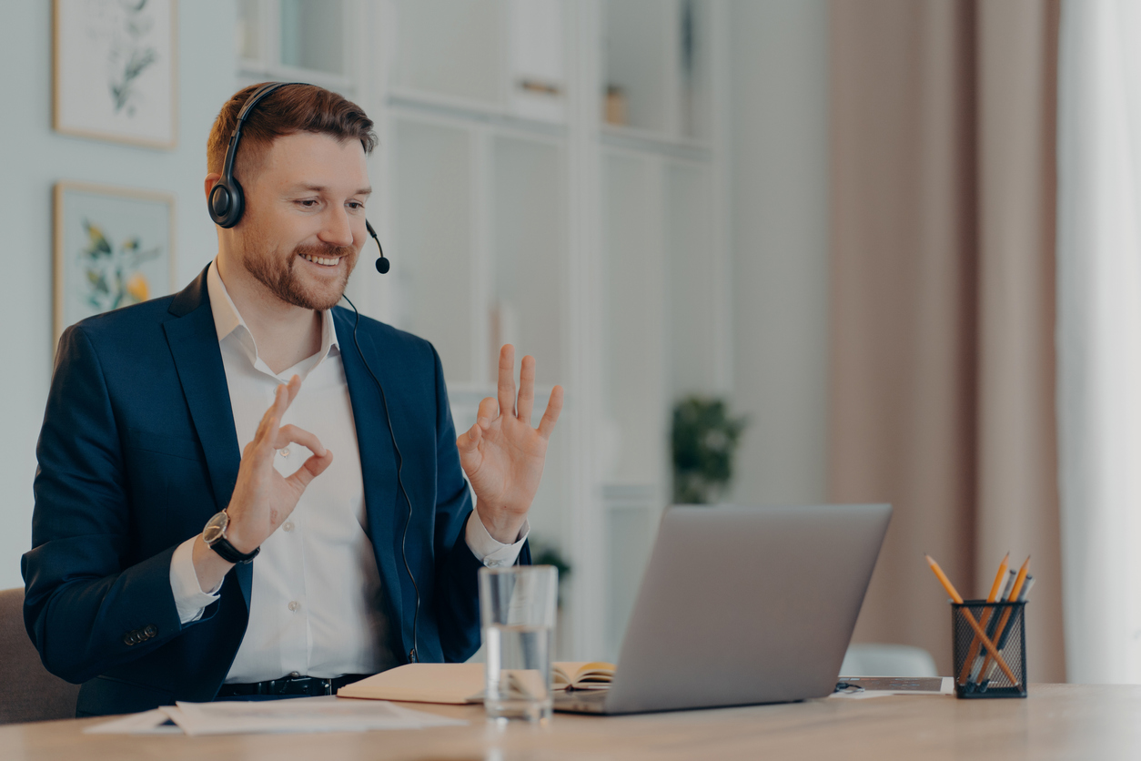 Career and distance job concept. Pleased bearded man corporate worker dressed formally gestures okay sign at webcam of laptop uses headset for online calling poses at desktop gains new knowledge