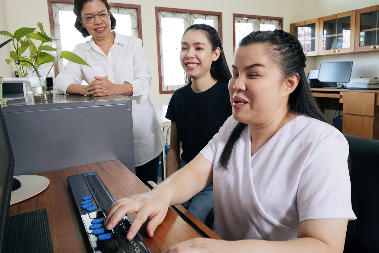 Colegas de trabalho mulheres asiáticas felizes em um ambiente de escritório, incluindo uma pessoa com deficiência visual que utiliza um computador com um dispositivo de leitura em brail