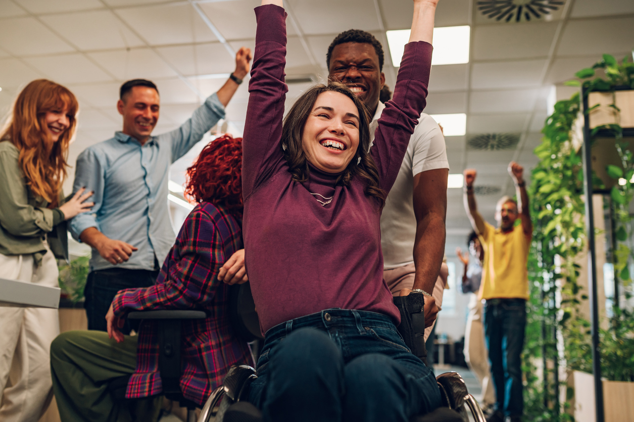 Diverse people having fun at work and racing on office chairs. Woman who uses wheelchair racing with a help of her african american colleague and winning the race. Everyone celebrating her success.